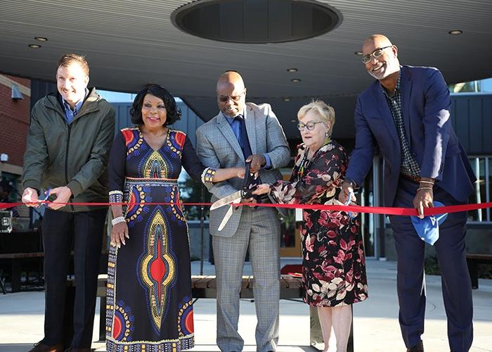 five people are behind a red ribbon, two have normal size scissors, three have their hands on a huge scissors